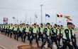 Agentes de policía en uniforme amarillo y negro marchan ordenadamente, con banderas de diferentes países ondeando de fondo.