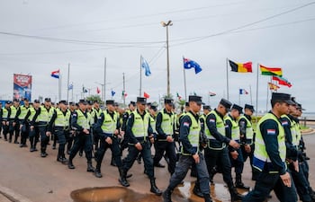 Agentes de policía en uniforme amarillo y negro marchan ordenadamente, con banderas de diferentes países ondeando de fondo.