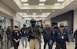 Hombre de cabello corto y barba, vestido con chaqueta negra, es llevado por dos policías armados con fusiles en un ambiente de aeropuerto.