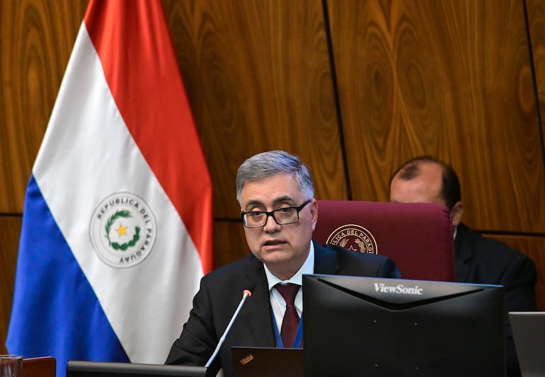 Hombre canoso con gafas y traje oscuro habla en reunión, acompañado por la bandera de Paraguay y una computadora ViewSonic.