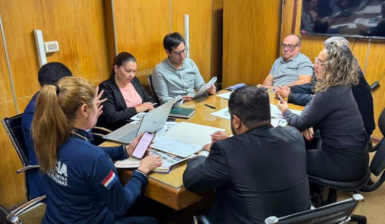 Siete personas en sala de reunión, un hombre gesticulando y una mujer con abrigo oscuro observando, documentos y laptop sobre la mesa.