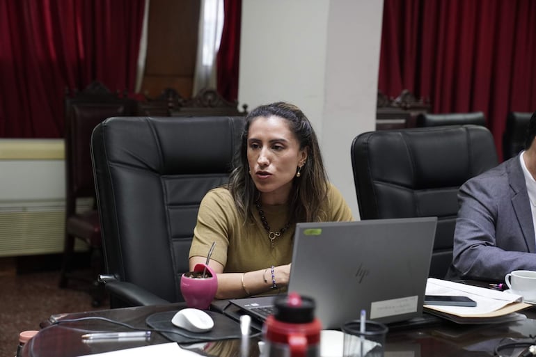 Bettina Albertini sentada en silla negra, habla con un hombre de saco gris en ambiente formal, con portátil y vaso de mate.
