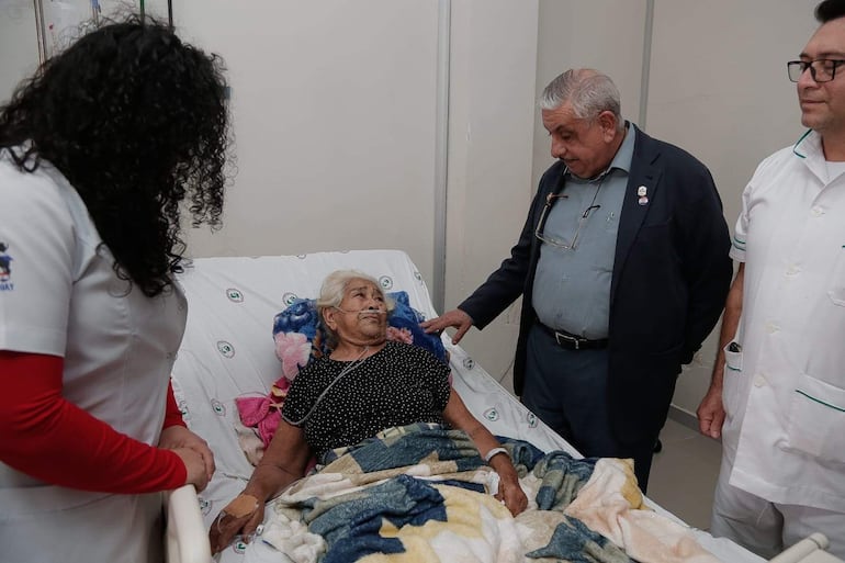 Mujer mayor en cama con tubo de oxígeno, rodeada de dos hombres, uno con gesto amable y médico observando, en ambiente hospitalario.