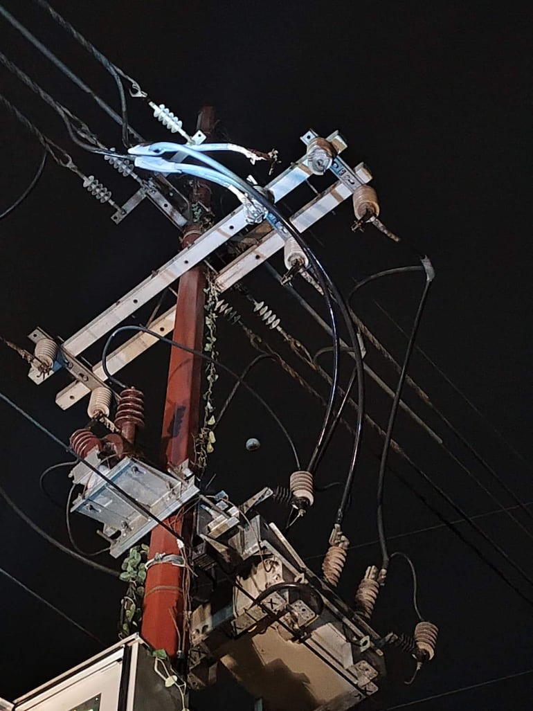 Poste de electricidad con cables y transformadores, rodeado de vegetación, en un ambiente nocturno.