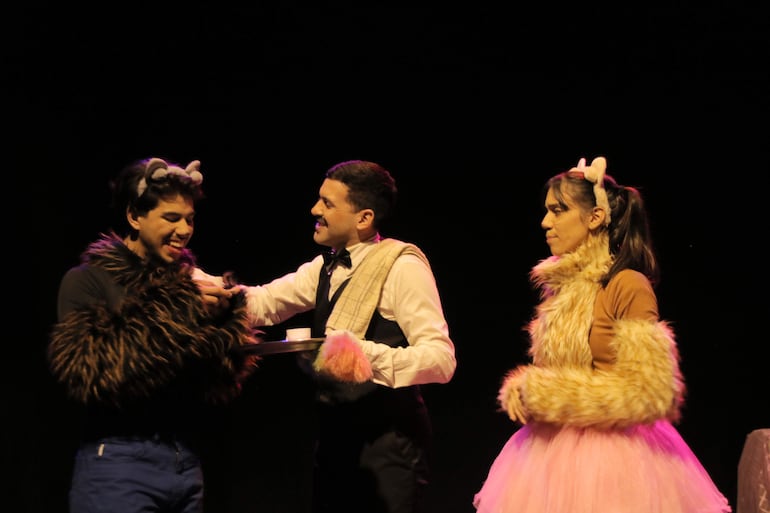 Hombre de camisa blanca con pajarita interactúa con disfraz peludo y mujer en falda de tul rosa, todos en un escenario iluminado.