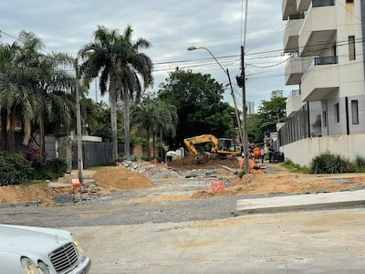 Tres trabajadores con chalecos naranjas rodean una excavadora amarilla en una calle con vegetación y vallas de seguridad.
