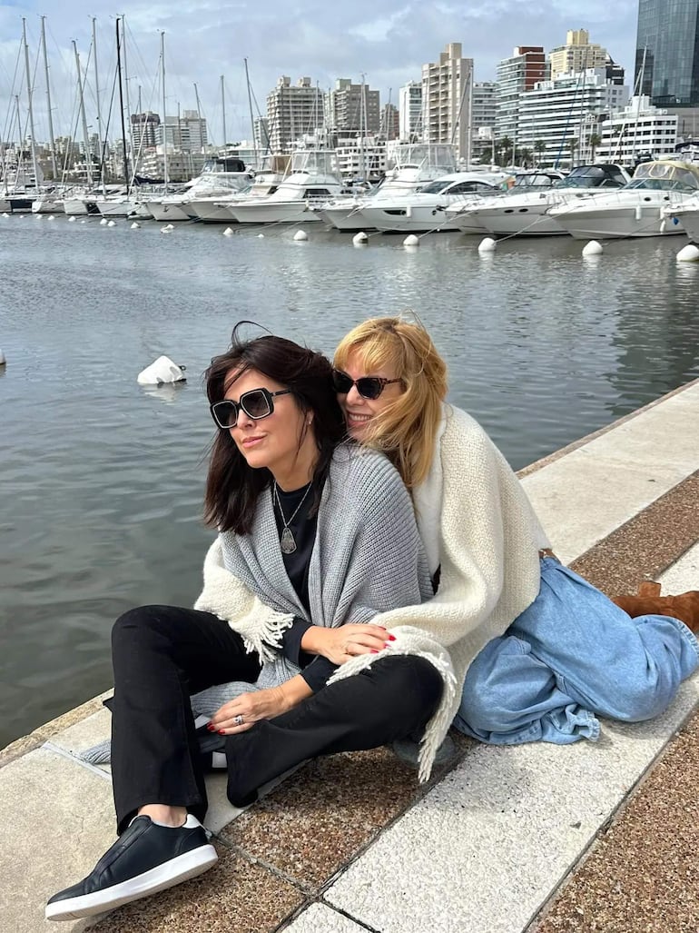 Paola Hermann con suéter gris y Vivian Benítez con suéter blanco, ambas con gafas de sol y sonrisas, sentadas junto a yates.