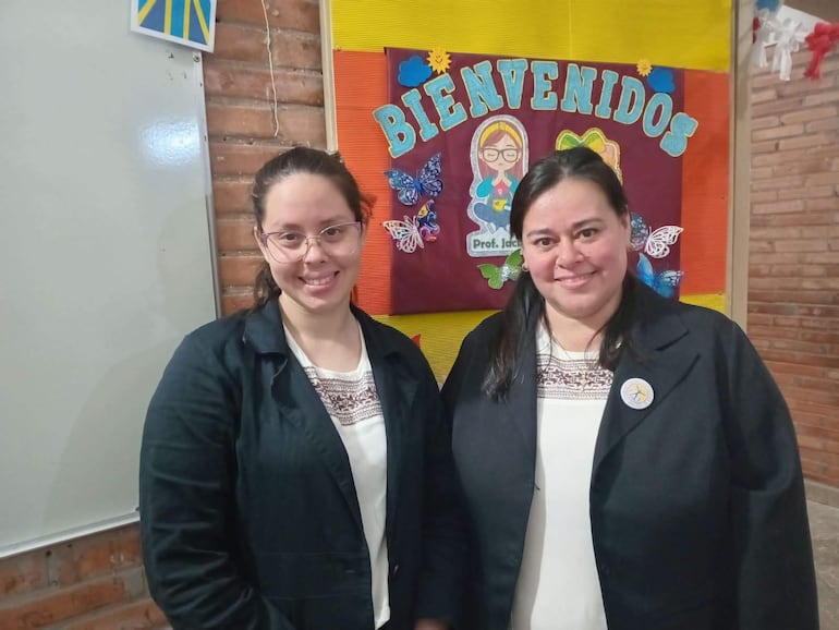 Lilian Giménez y Jacqueline Jaquet sonríen en un aula, vestidas con chaquetas y blusas claras, ante un cartel colorido que dice 'BIENVENIDOS'.