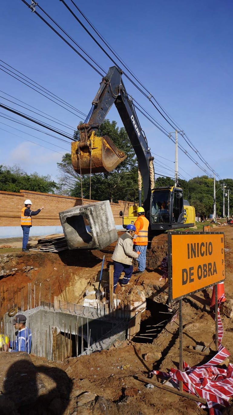 Trabajador con chaleco naranja y casco da instrucciones, mientras operador trabaja en una excavadora amarilla en la obra de construcción.