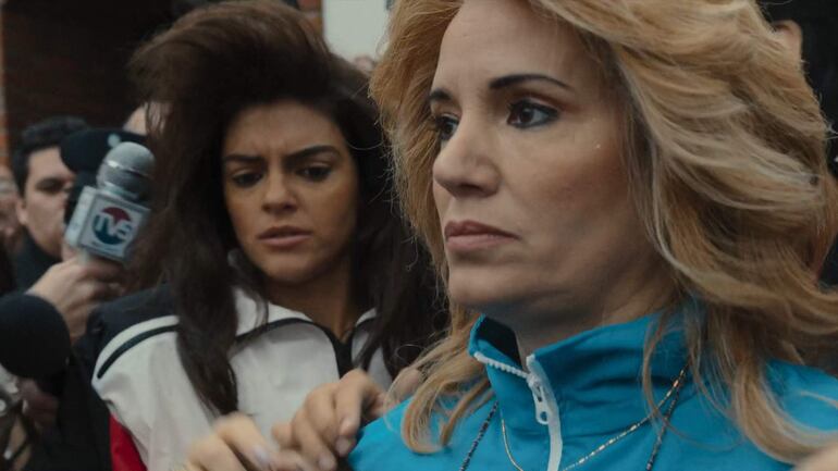 Mujeres en primer plano: una con chaqueta azul y expresión seria, la otra con cabello rizado; hombres y micrófono de fondo.
