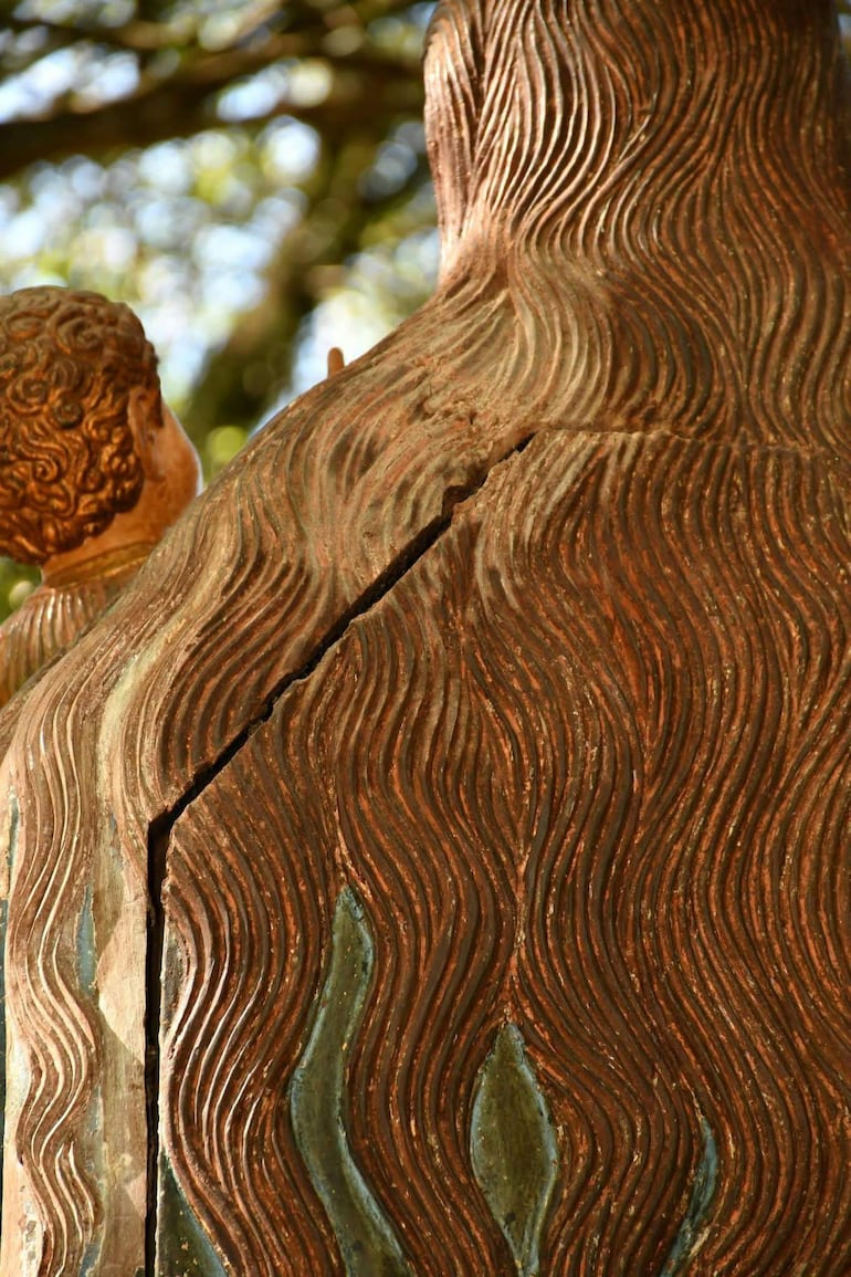 Escultura de una figura humana mayor con cabello ondulado junto a un niño, en un ambiente natural con vegetación difusa al fondo.
