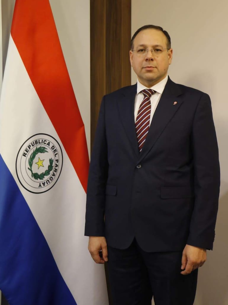 Adolfo Vallejos en traje oscuro y corbata a rayas, de pie frente a la bandera de Paraguay en un ambiente formal.