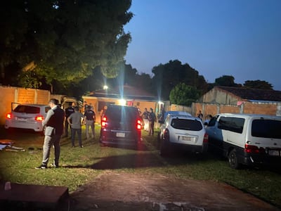 Hombre con traje negro revisa su teléfono mientras agentes de policía realizan un allanamiento en un patio oscuro.