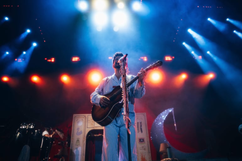 Músico en pijama a rayas sostiene guitarra en escenario iluminado con luces azul y naranja, creando ambiente festivo.