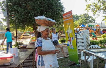 Mujer mayor sonriente con canasta en la cabeza, rodeada de otras personas en un ambiente festivo y comunitario.