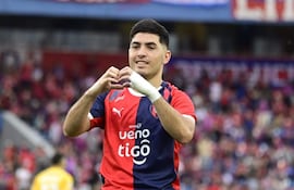 Ignacio Aliseda sonriente con camiseta roja y azul, brazos formando un corazón, en un estadio lleno de aficionados animados.