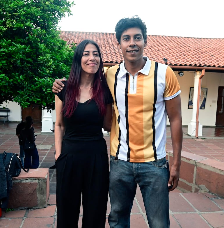Mujer de cabello largo con camisa negra y hombre en camiseta colorida sonríen entrelazados en un ambiente cultural al aire libre.