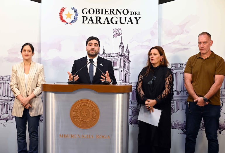 Eduardo Gustale en traje frente a micrófono, Adriana Ortiz Semidei en chaqueta de rayas, ambiente institucional en Palacio de Gobierno.