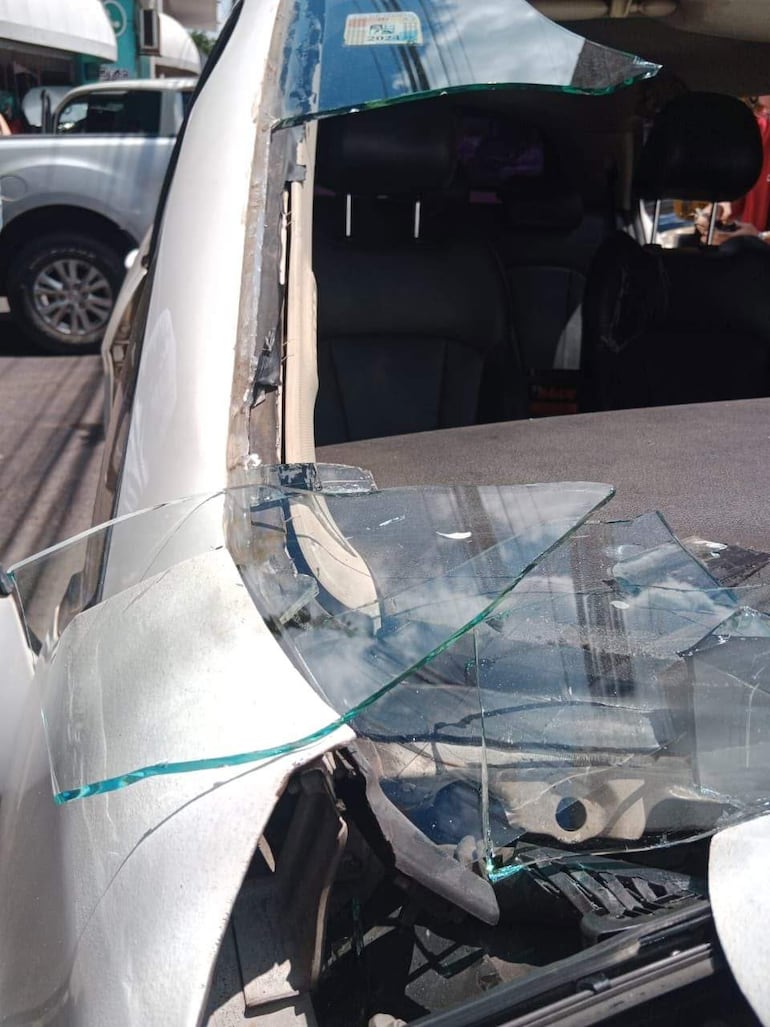 Coche con puerta dañada y fragmentos de vidrio esparcidos, en una calle urbana con otros vehículos y peatones al fondo.