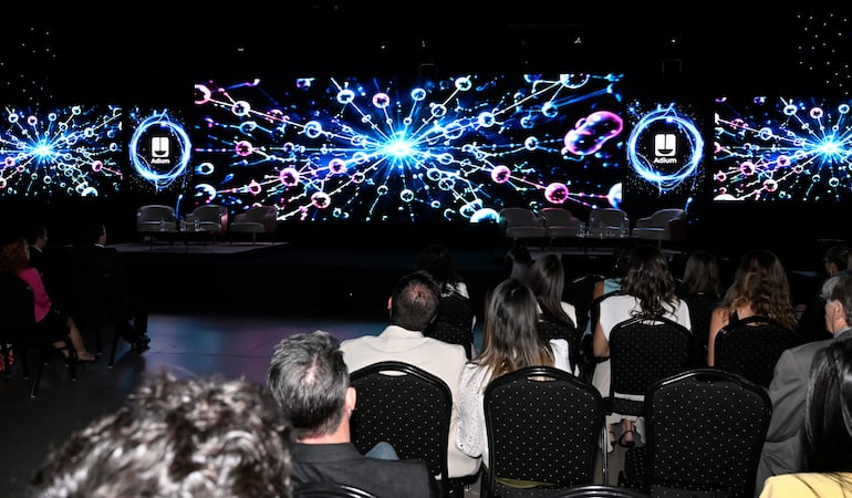 Audiencia sentada en filas en un evento, con luces tenues y un escenario colorido exhibiendo el logo de Adlum.
