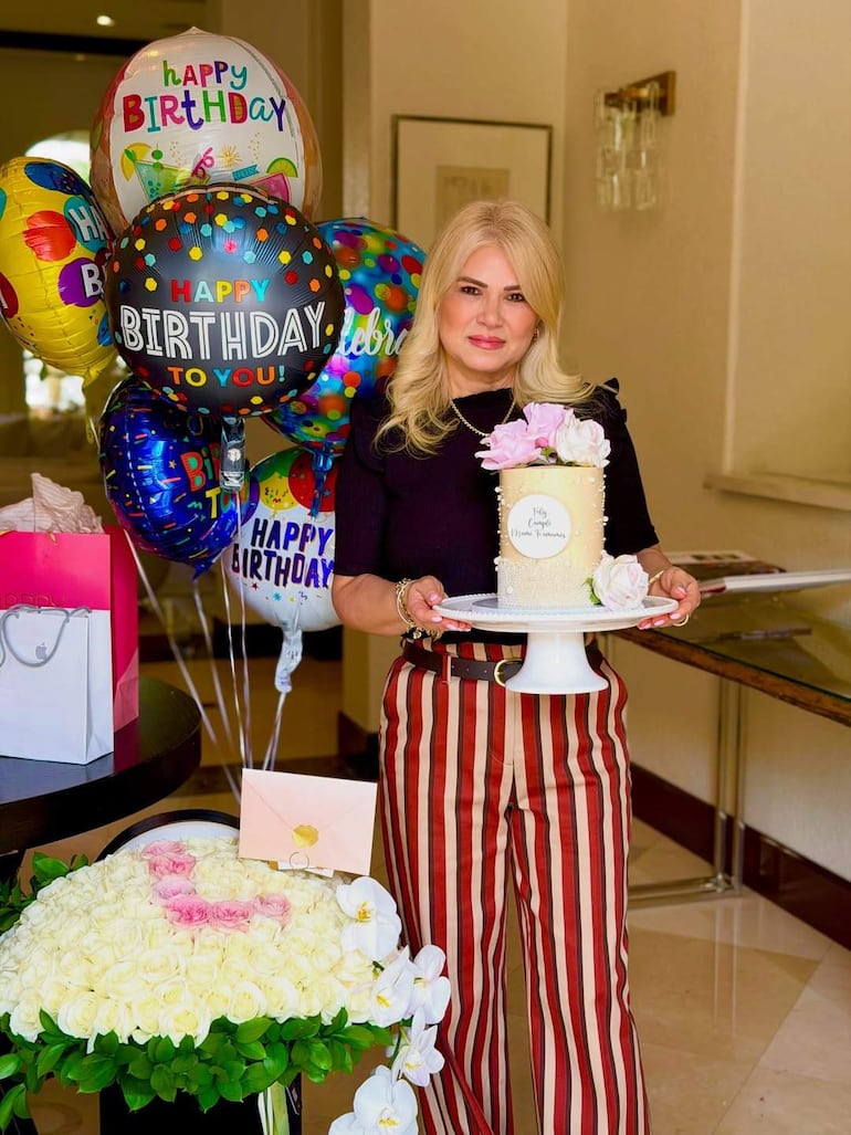 Mujer rubia en camiseta negra y pantalones a rayas sostiene un pastel con flores, rodeada de globos coloridos.
