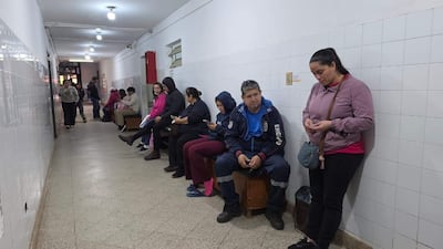 Varias personas en un pasillo del hospital, esperando atención; una mujer con chaqueta rosa observa su teléfono.