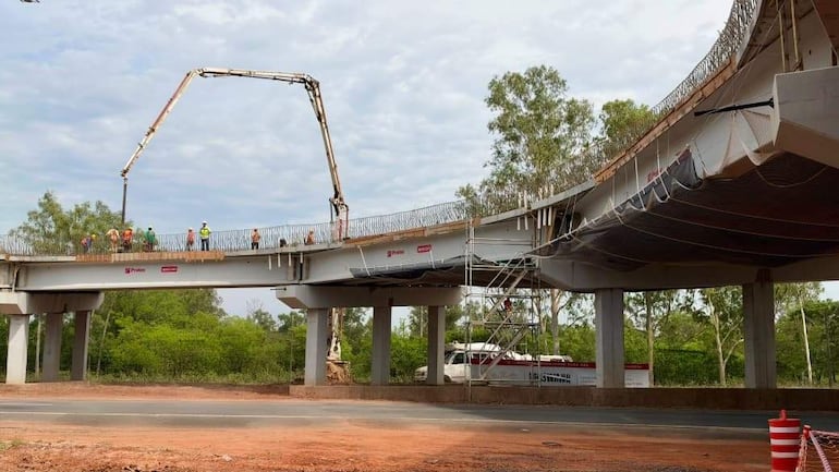 Trabajadores en chalecos de seguridad y cascos, usando un brazo mecánico para verter concreto en el viaducto en construcción.