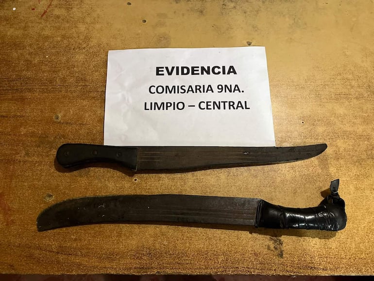 Cuchillos y machetes sobre superficie de madera con documento que dice 'EVIDENCIA' en un entorno policial.