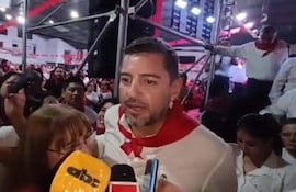 Hombre de camisa blanca y pañuelo rojo conversa con mujer con micrófono, rodeados de asistentes entusiastas en un ambiente político.