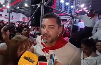 Hombre de camisa blanca y pañuelo rojo conversa con mujer con micrófono, rodeados de asistentes entusiastas en un ambiente político.