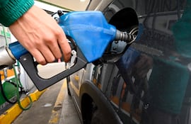 Mano sujeta bomba de combustible en estación de servicio, prenda verde visible, ambiente cerrado con bombas al fondo.