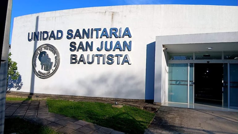 Fachada blanca de la Unidad Sanitaria San Juan Bautista, con logo y nombre visible en letras metálicas, rodeada de área verde.
