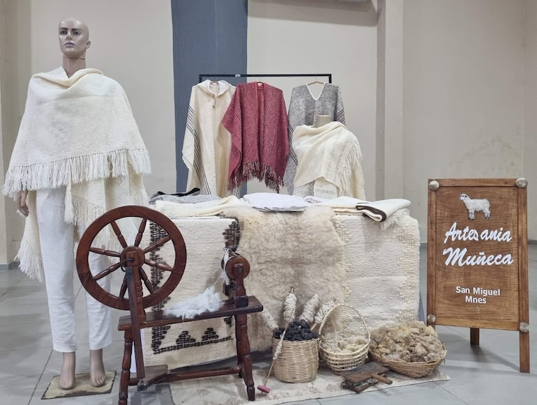Un maniquí viste un poncho blanco con flecos, rodeado de textiles coloridos y un huso sobre la mesa en el stand de Artesanía Muñeca.