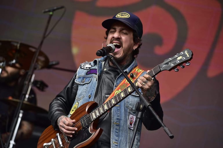 Músico en chaqueta de mezclilla con gorra y guitarra, cantando apasionadamente en el escenario al aire libre.