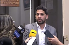 Raúl Benítez en traje gris claro, rodeado de reporteros con micrófonos, en la Comisión de Presupuesto con inscripción detrás.