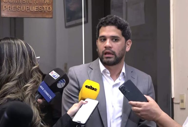 Raúl Benítez en traje gris claro, rodeado de reporteros con micrófonos, en la Comisión de Presupuesto con inscripción detrás.