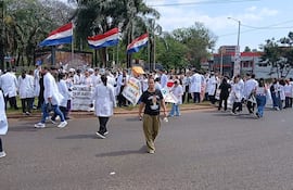 Mujer en camiseta negra camina entre médicos con batas blancas y pancartas en manifestación exterior, árboles y edificios al fondo.