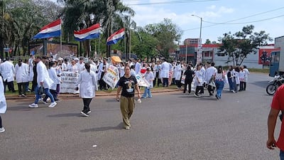 Mujer en camiseta negra camina entre médicos con batas blancas y pancartas en manifestación exterior, árboles y edificios al fondo.