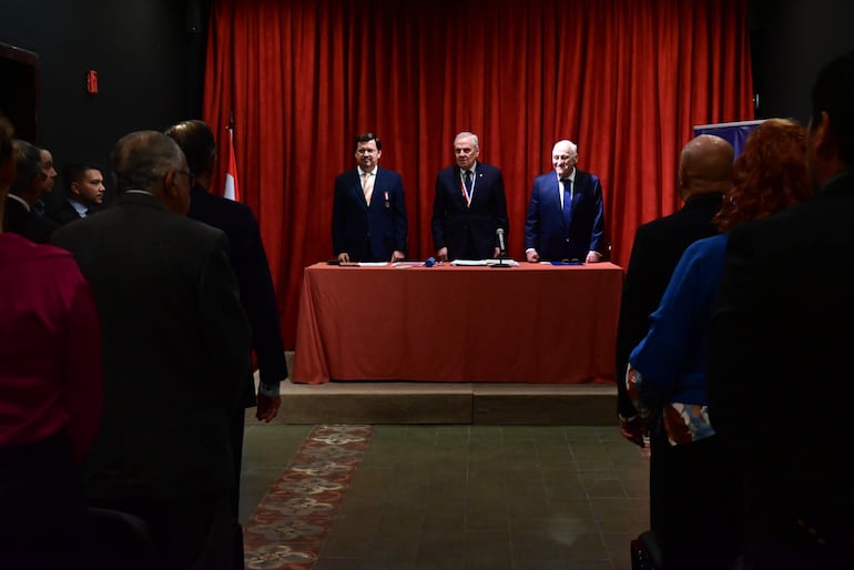 Mujer con abrigo oscuro y vestido claro junto a hombre de traje gris y corbata roja, en ambiente formal conversando.