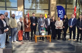 Grupo de 12 personas, hombres y mujeres en trajes formales, sonríen en un evento al aire libre con banderas de fondo.