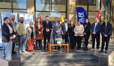 Grupo de 12 personas, hombres y mujeres en trajes formales, sonríen en un evento al aire libre con banderas de fondo.