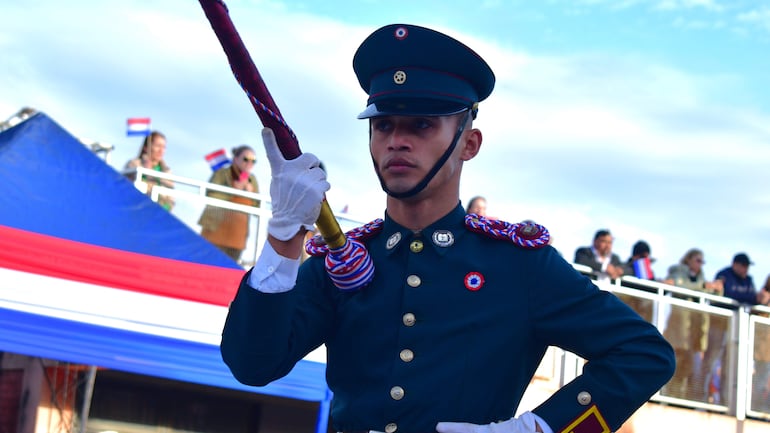 Joven militar en uniforme verde oscuro con detalles rojos sostiene un bastón, en un ambiente colorido con banderas paraguayas.