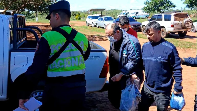 Ismael Obregón Olmedo rodeado de hombres y un policía, en un ambiente rural y soleado, dirigiéndose a un coche policía.