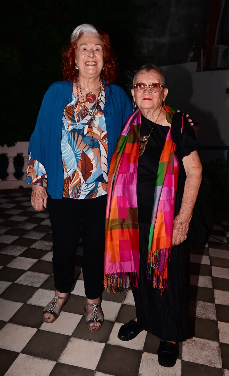 Lucy Yegros con gafas oscuras y vestido negro sonríe mientras conversa con otra mujer de blusa colorida y chaqueta azul.