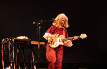 Músico con cabello largo y camisa blanca, tocando guitarra en un escenario oscuro, concentrado en su interpretación.