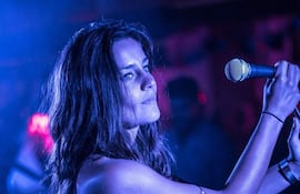 Kelly Ou Moi canta sosteniendo un micrófono en ambiente tenue de luces azules y anaranjadas, con el público en el fondo.
