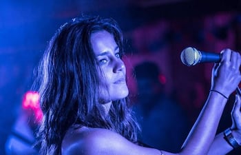 Kelly Ou Moi canta sosteniendo un micrófono en ambiente tenue de luces azules y anaranjadas, con el público en el fondo.