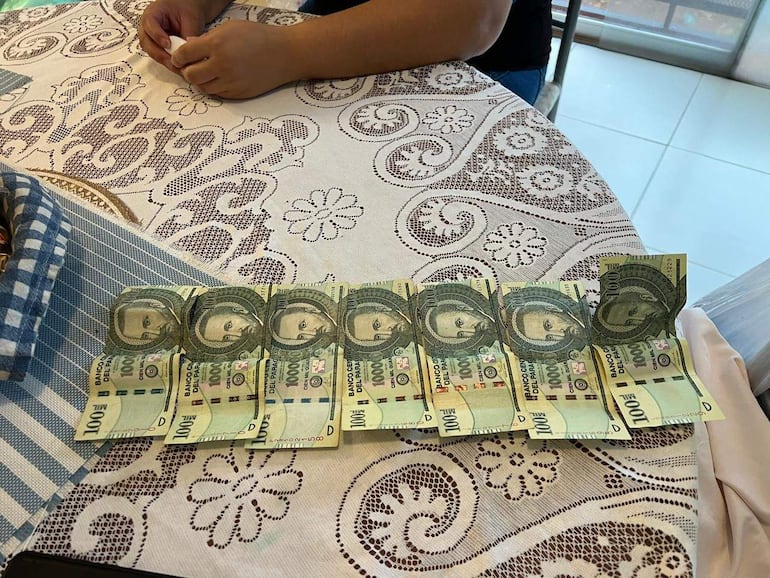 Billetes de 100.000 guaraníes organizados sobre una mesa con mantel floral. Mano sosteniendo un objeto pequeño.