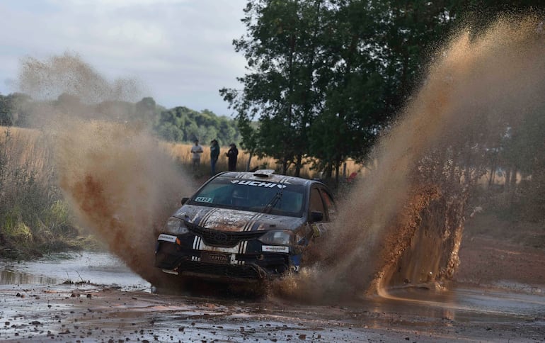 Automóvil de rally salpicando barro en un entorno natural, con espectadores difusos en el fondo.