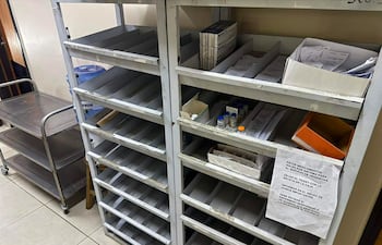 Estanterías metálicas en un área de almacenamiento médica, con cartel sobre entrega de medicamentos y un carrito metálico visible.